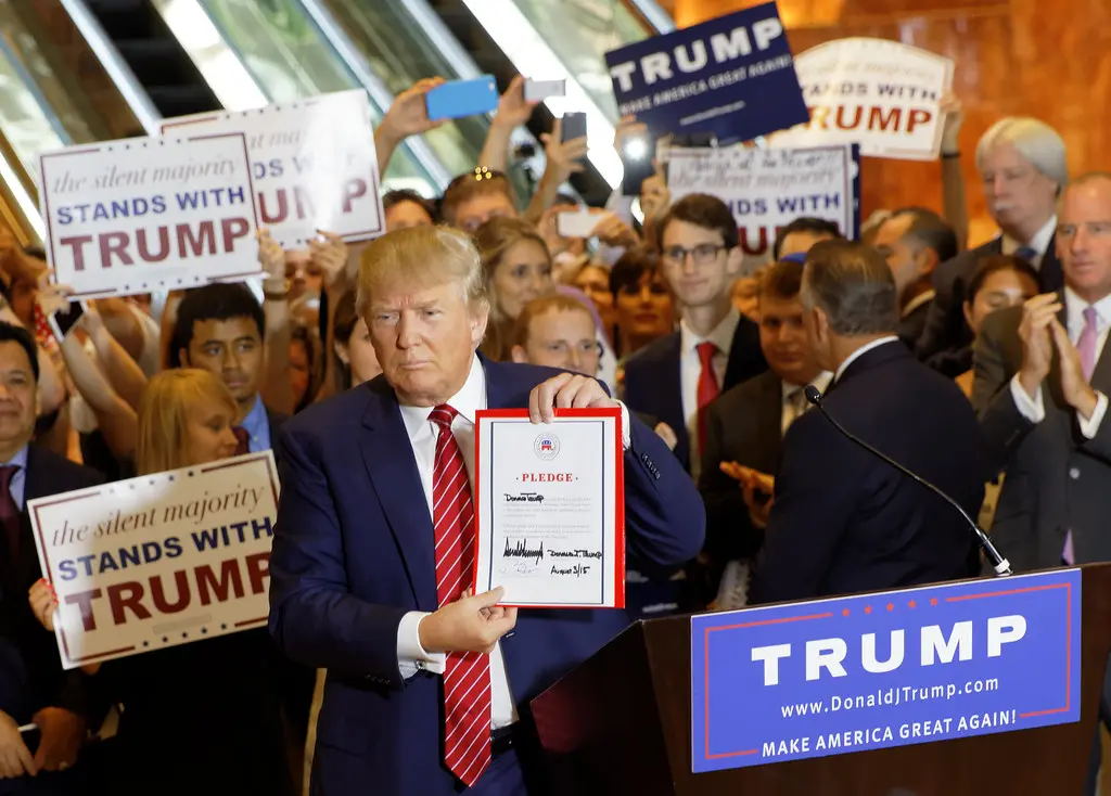 Donald Trump holding up a signed pledge document while standing at a podium surrounded by supporters.